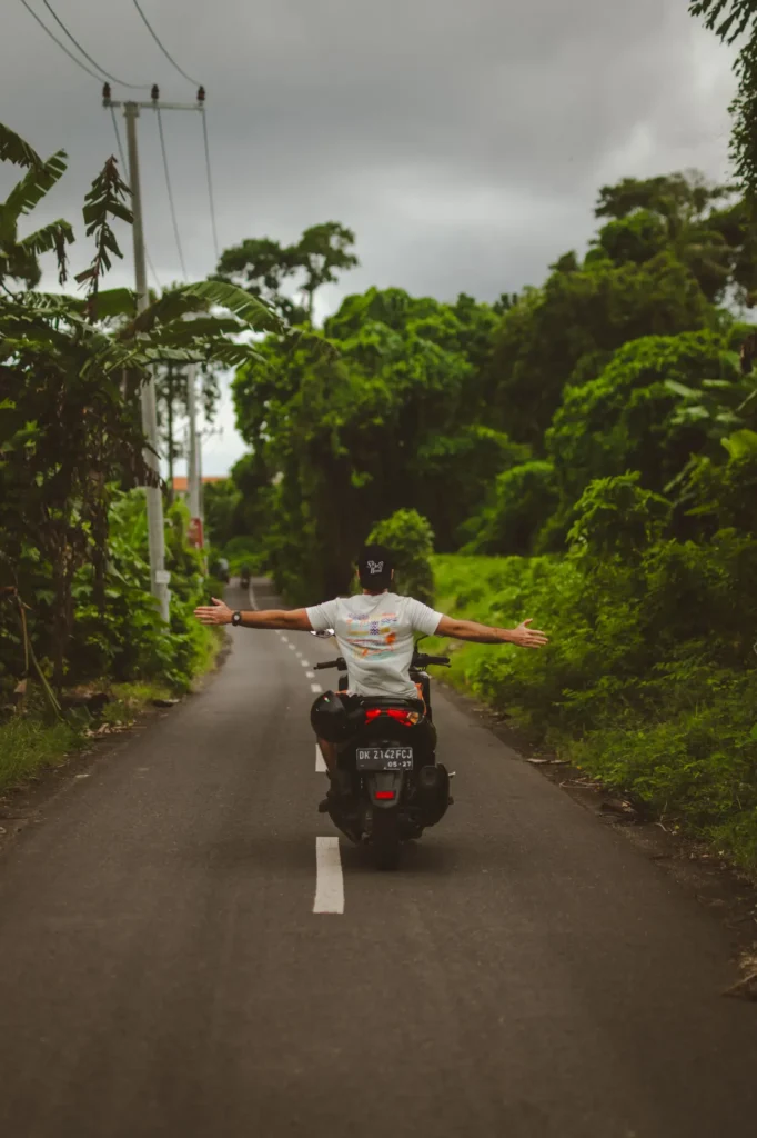 A rider seen from behind with arms outstretched on an empty road lined with jungle in Bali