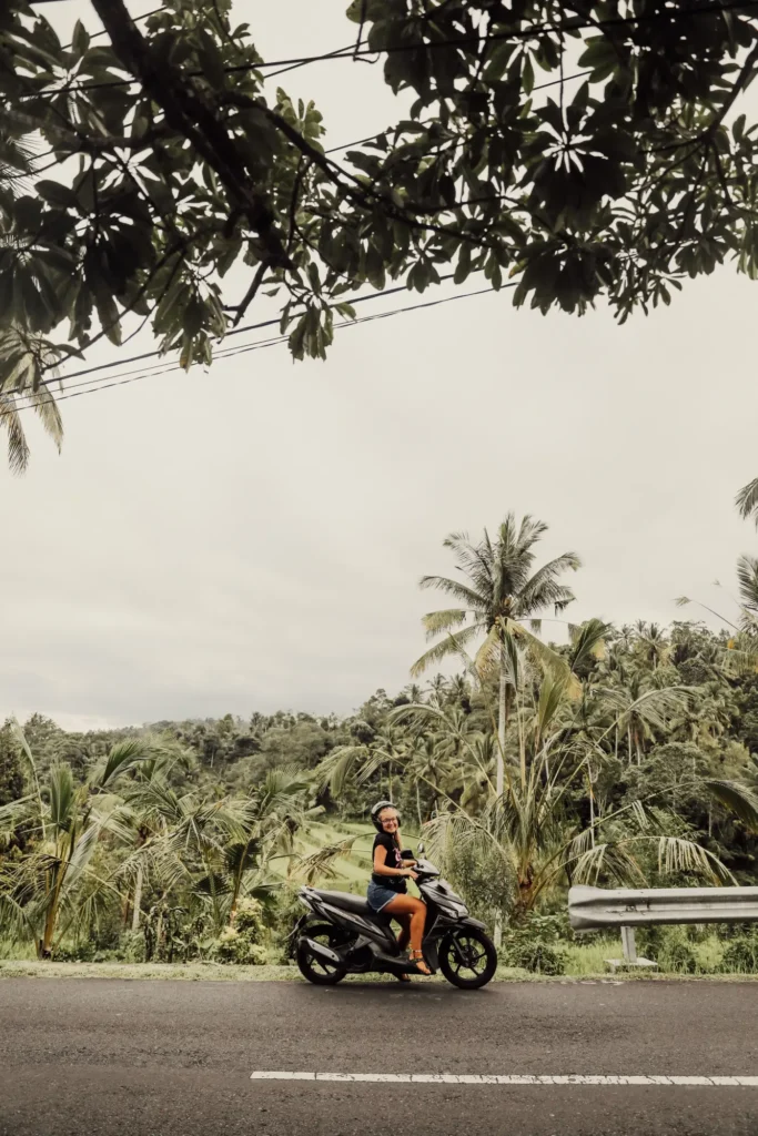 A rider on a scooter on an open road below coconut palms under an overcast sky in Bali