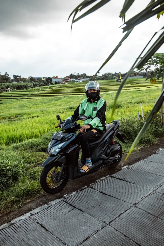 A rider in a green jacket on a black scooter on a path beside rice terraces in Bali