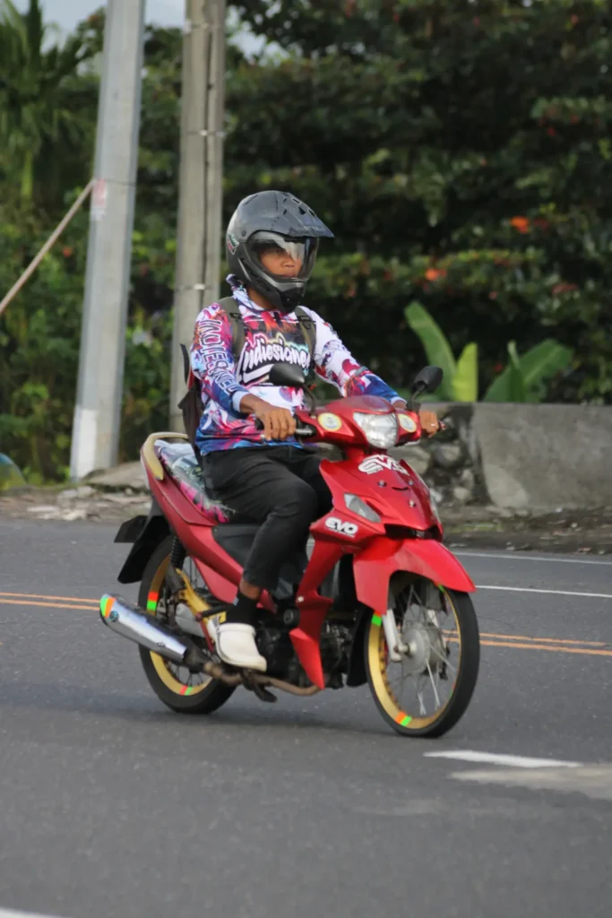 A rider in a patterned jacket on a red underbone motorbike on a main road in Bali