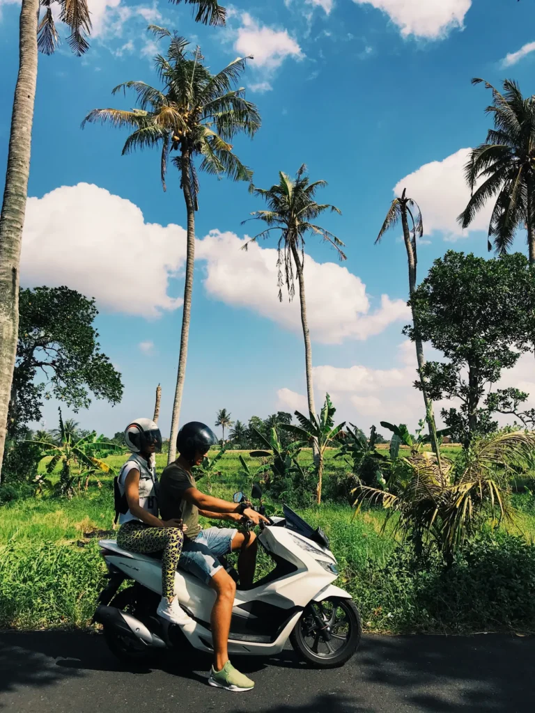 Two people on a white maxi-scooter beside rice fields and coconut palms in Bali